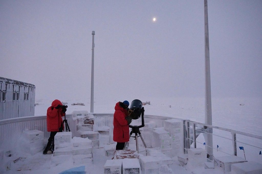 Two people bundled up in thick red winter jackets stand outside next to two telescopes. Everything around them is coated in white snow.